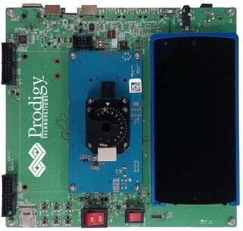 A green Prodigy Technovations printed circuit board (PCB) featuring a central blue test module with a black rotary dial and a smartphone attached to the right side.