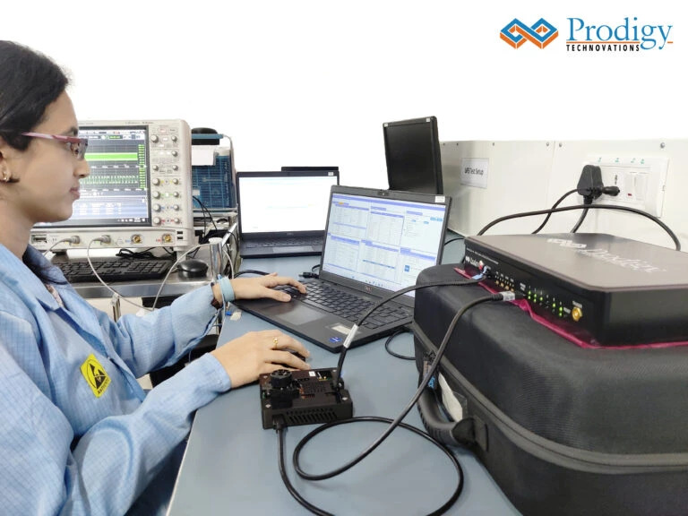 An engineer in a blue lab coat sits at a desk using a laptop and a Tektronix oscilloscope to analyze data from a Prodigy Technovations Automotive Ethernet Protocol Analyzer connected to testing hardware.